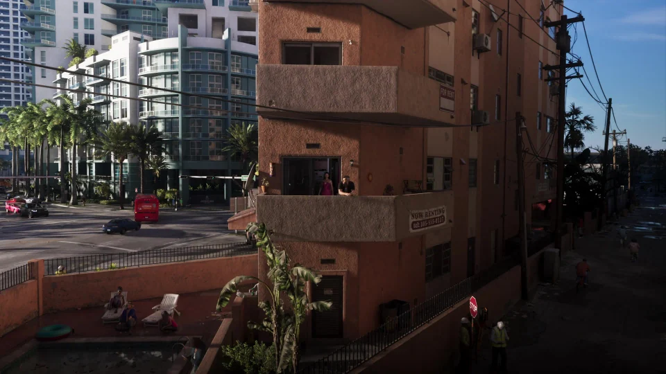 Jason und Lucia auf dem Balkon eines rosa-orangen Gebäudes neben einer Straße und Hochhäusern